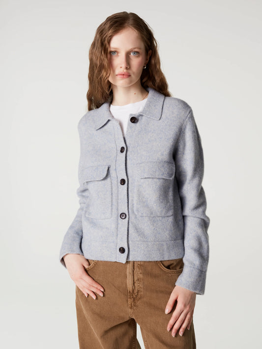 A woman modeling a light gray cropped button-up cardigan with flap pockets over a white t-shirt and brown corduroy pants, shot against a plain light gray studio background.