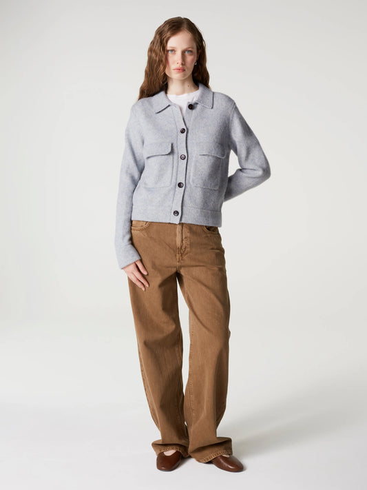 A model stands against a light gray studio background wearing a light gray cropped button-front cardigan over a white top, paired with brown wide-leg pants and brown shoes.