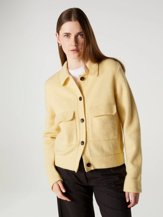 A woman modeling a pale yellow textured button-up jacket with dark buttons, worn over a white top and black trousers, against a light gray studio background.