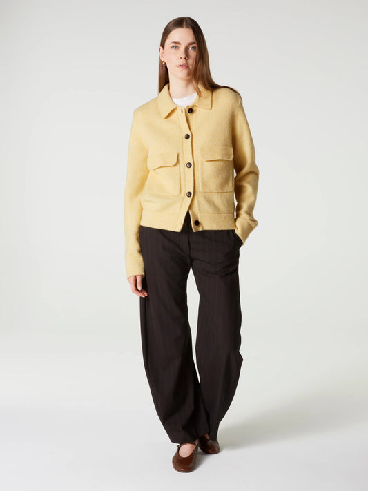A model stands against a light gray studio background wearing a pale yellow cropped button-front jacket over a white top, dark brown wide-leg trousers, and brown shoes.