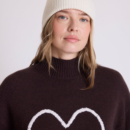 A woman wearing a cream ribbed knit beanie and a dark brown turtleneck sweater with a white heart outline on the front, photographed against a plain light gray background.