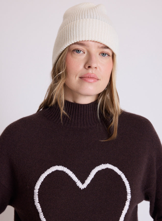 A woman wearing a cream ribbed knit beanie and a dark brown turtleneck sweater with a white heart outline on the front, photographed against a plain light gray background.