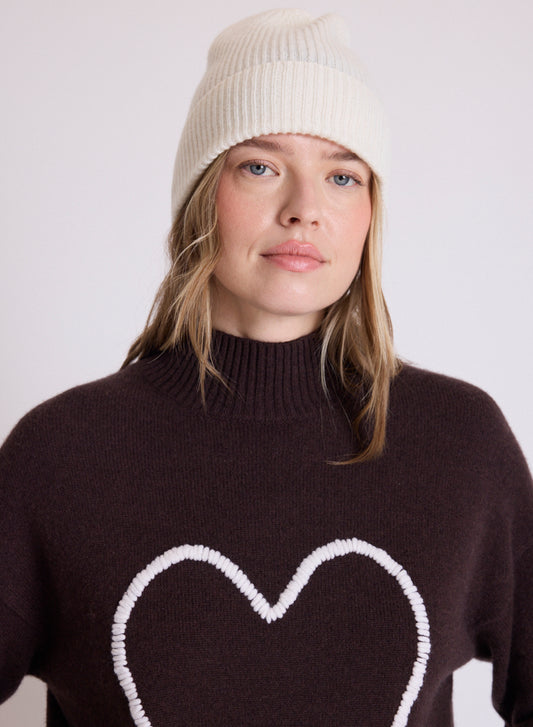 A woman wearing a cream ribbed knit beanie and a dark brown turtleneck sweater with a white heart outline on the front, photographed against a plain light gray background.