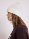 A woman in profile wearing a ribbed cream beanie and a dark brown sweater, photographed against a plain light background.