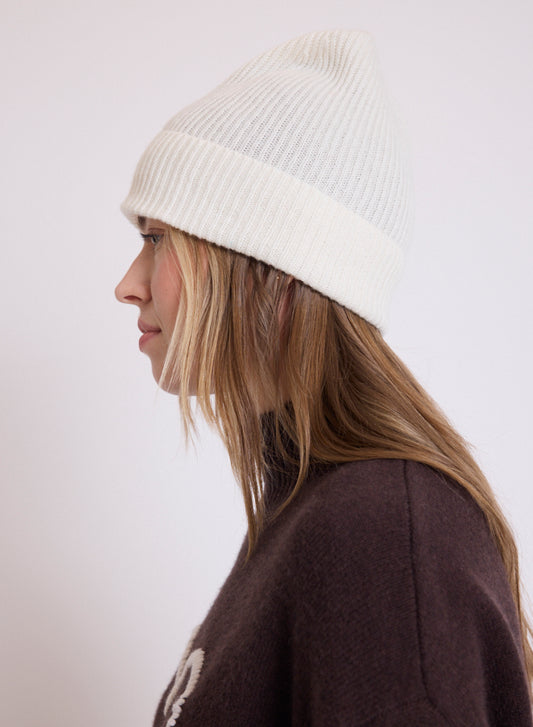 A woman in profile wearing a ribbed cream beanie and a dark brown sweater, photographed against a plain light background.