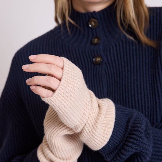 A close-up of a person wearing a navy ribbed knit mock-neck sweater with dark buttons and cream ribbed knit fingerless wrist cuffs.