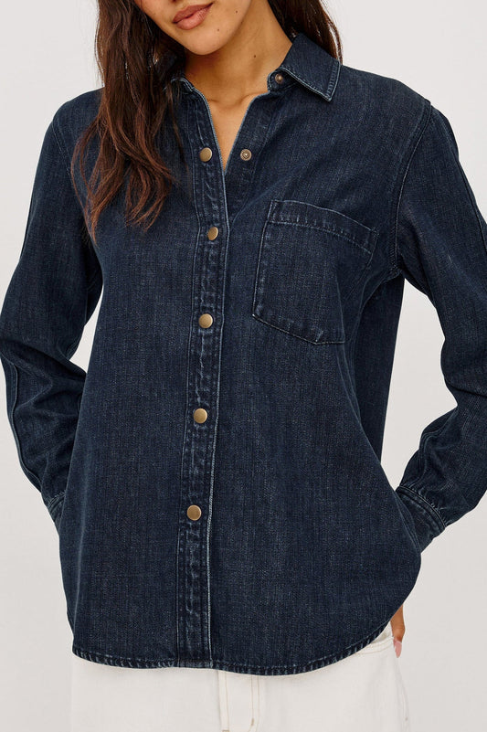 A woman wearing a dark indigo denim long-sleeve button-up shirt with a chest pocket, paired with white pants, shown against a plain light background.