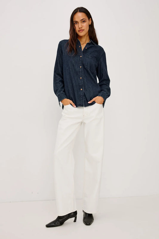 A woman modeling a dark indigo denim long-sleeve button-up shirt paired with white wide-leg pants and black pointed-toe heels, standing against a plain white studio background.