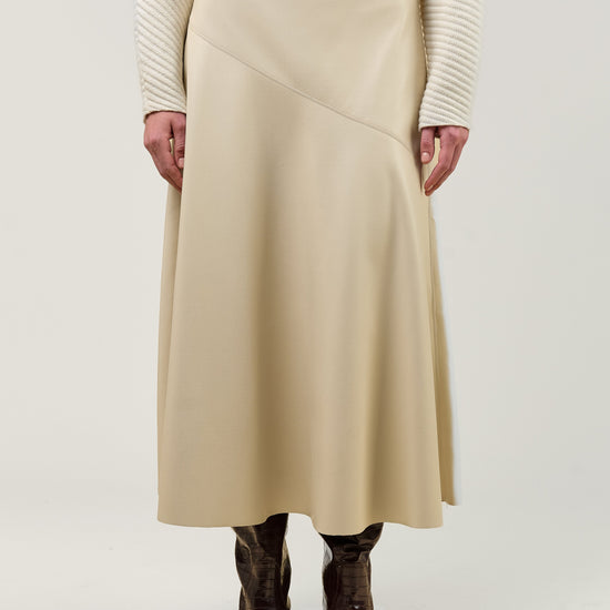A model is shown from the chest down wearing a beige wrap-style midi skirt, a cream ribbed sweater, and dark brown boots, set against a plain light gray studio background.