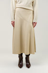 A model is shown from the chest down wearing a beige wrap-style midi skirt, a cream ribbed sweater, and dark brown boots, set against a plain light gray studio background.