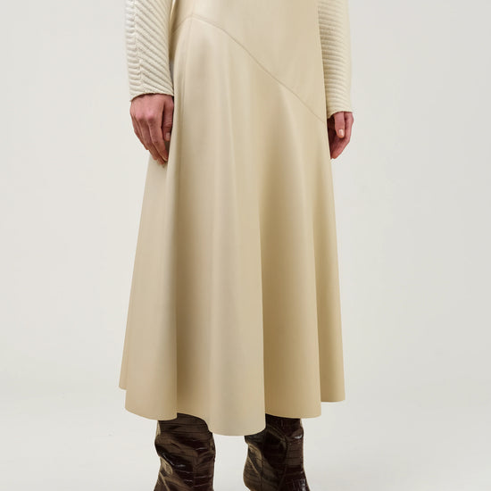 A model wearing a cream-colored pleated midi skirt, a cream ribbed sweater, and dark brown pointed-toe heeled boots, shown from the waist down against a light gray studio background.