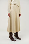 A model wearing a cream-colored pleated midi skirt, a cream ribbed sweater, and dark brown pointed-toe heeled boots, shown from the waist down against a light gray studio background.