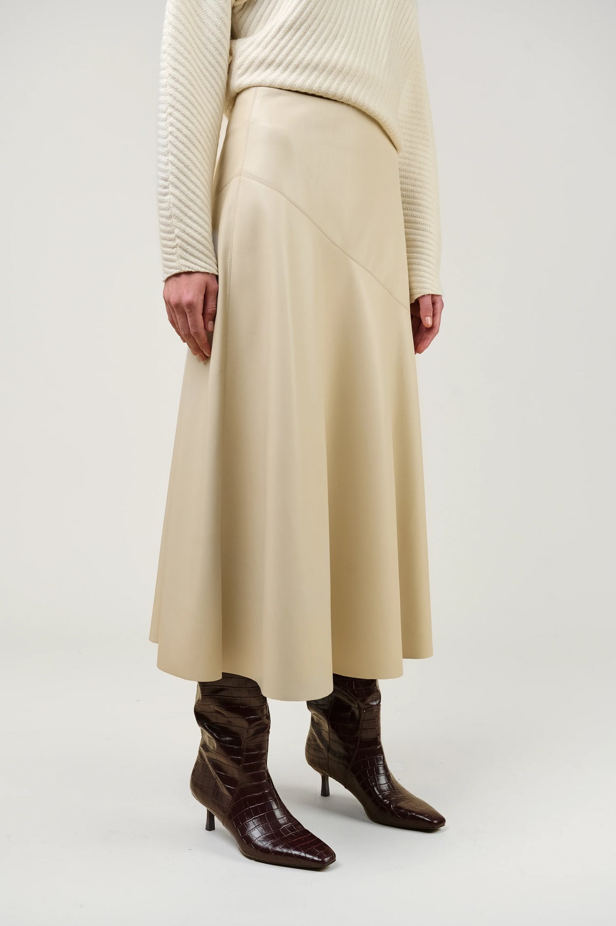 A model wearing a cream-colored pleated midi skirt, a cream ribbed sweater, and dark brown pointed-toe heeled boots, shown from the waist down against a light gray studio background.