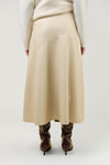 A person shown from the back wearing a long cream ribbed sweater, a beige wrap-style midi skirt, and brown heeled ankle boots against a light gray studio background.