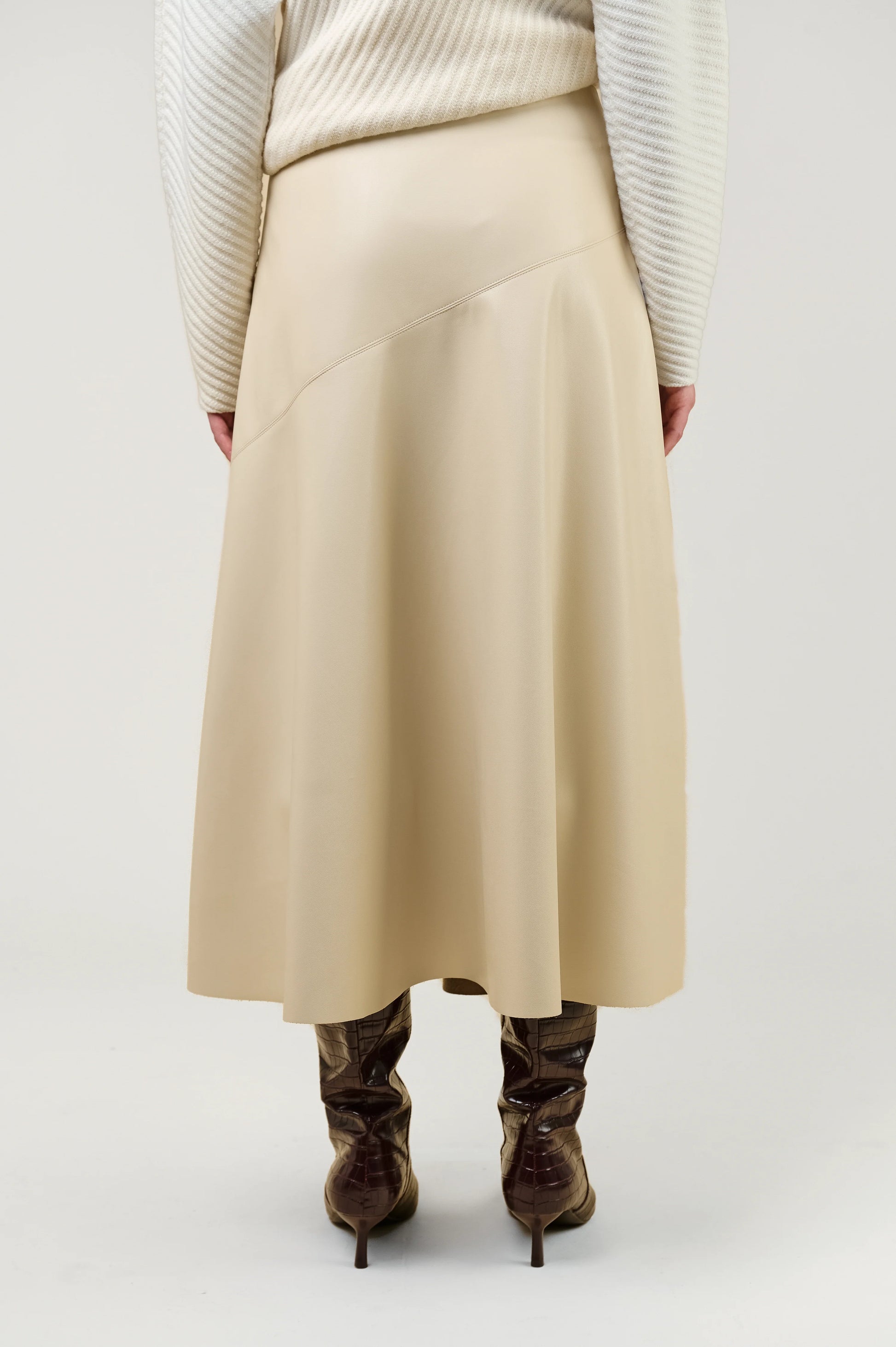 A person shown from the back wearing a long cream ribbed sweater, a beige wrap-style midi skirt, and brown heeled ankle boots against a light gray studio background.