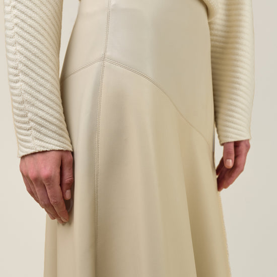 Close-up of a person wearing a cream ribbed knit sweater and a matching cream high-waisted skirt with smooth paneling and a draped waist seam, photographed against a plain light beige background.