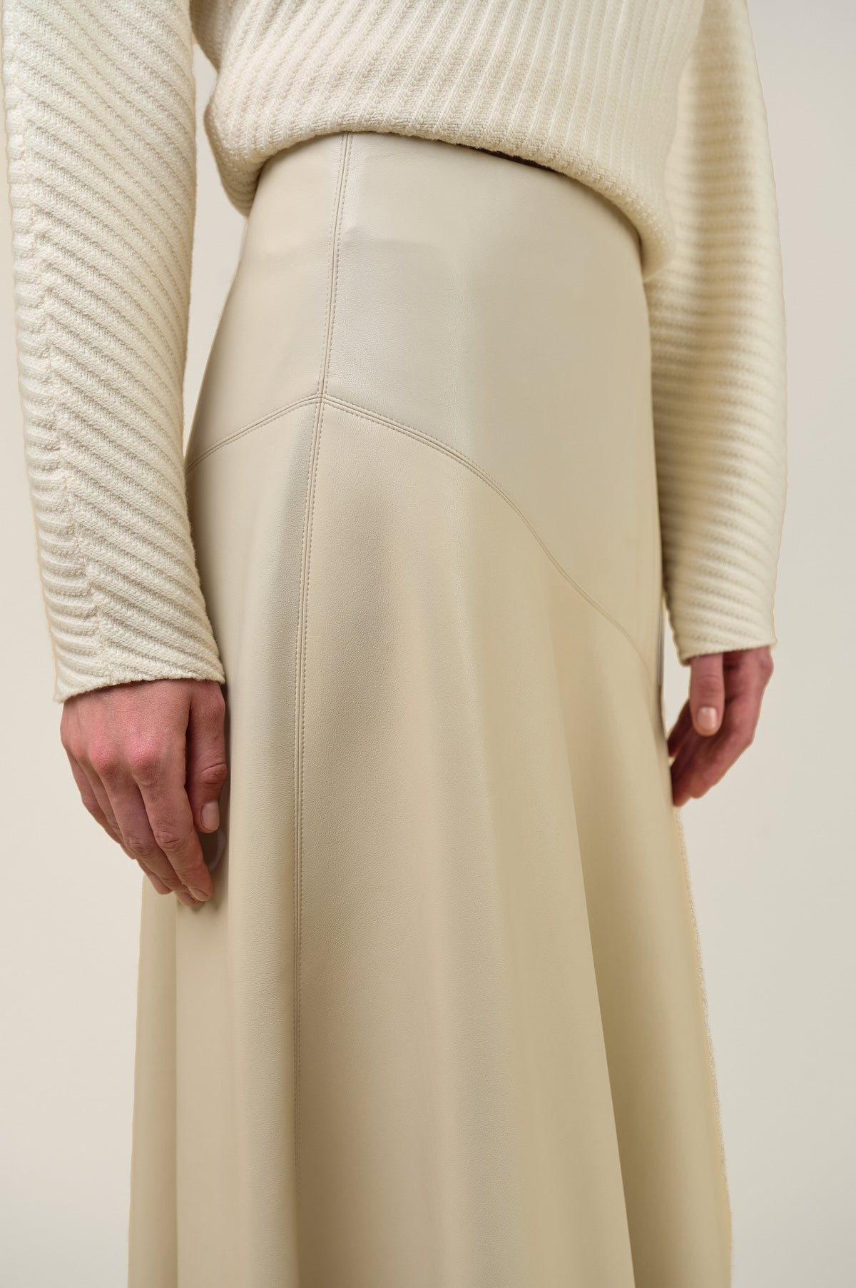 Close-up of a person wearing a cream ribbed knit sweater and a matching cream high-waisted skirt with smooth paneling and a draped waist seam, photographed against a plain light beige background.