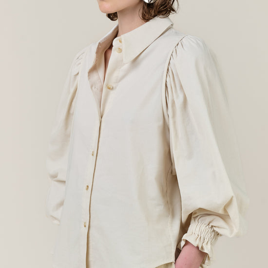 A model wearing a cream-colored long-sleeve button-up shirt with dramatic puffed balloon sleeves and gathered cuffs, paired with beige corduroy trousers, against a solid light beige background.