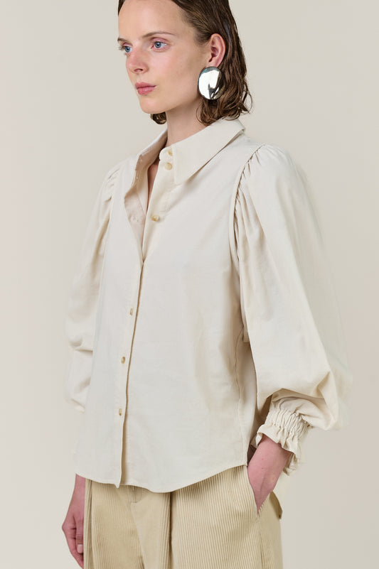 A model wearing a cream-colored long-sleeve button-up shirt with dramatic puffed balloon sleeves and gathered cuffs, paired with beige corduroy trousers, against a solid light beige background.