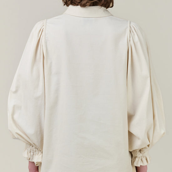 Back view of a model wearing a cream-colored blouse with dramatic balloon sleeves and gathered cuffs, paired with light beige trousers, against a solid light beige background.