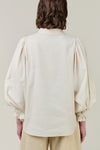Back view of a model wearing a cream-colored blouse with dramatic balloon sleeves and gathered cuffs, paired with light beige trousers, against a solid light beige background.