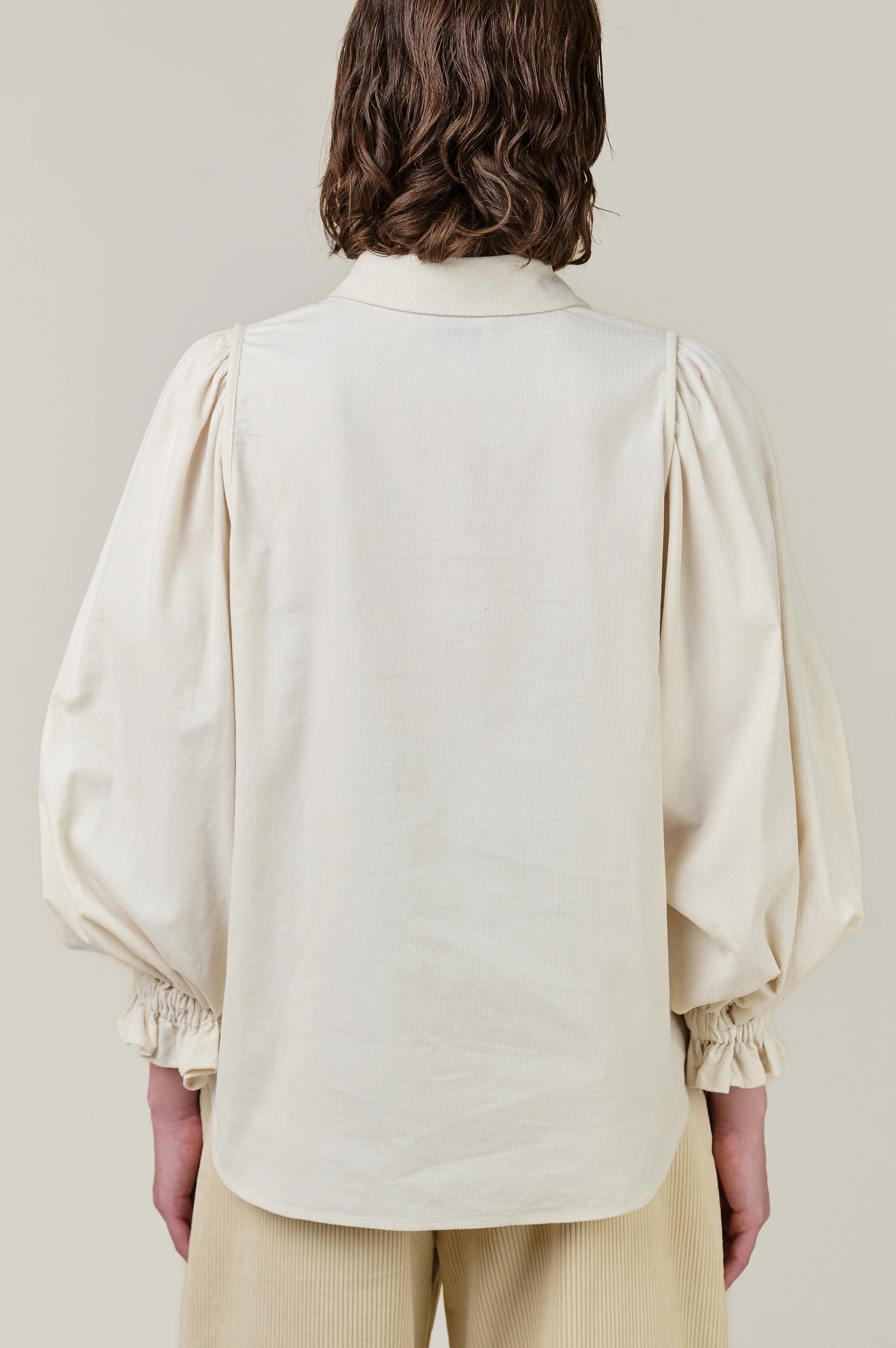 Back view of a model wearing a cream-colored blouse with dramatic balloon sleeves and gathered cuffs, paired with light beige trousers, against a solid light beige background.