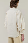 A model shown from the back wearing a cream-colored blouse with dramatic puffed balloon sleeves and gathered ruffled cuffs, paired with beige trousers, against a solid light beige background.