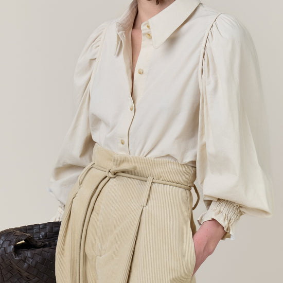 A model shown in profile wearing a cream-colored blouse with dramatic puffed sleeves and a matching beige corduroy skirt with a tie waist. She is also wearing a large, reflective silver earring and holding a dark woven handbag against a solid light beige background.