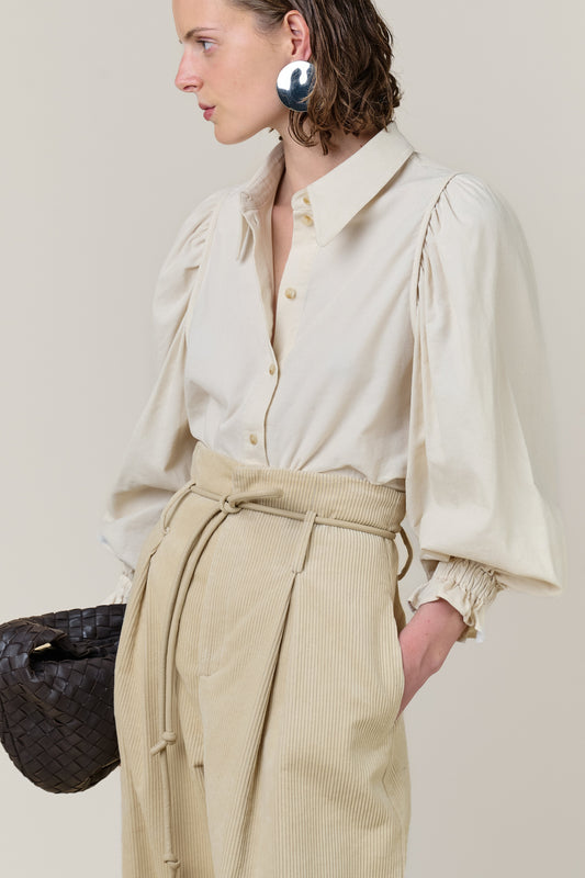 A model shown in profile wearing a cream-colored blouse with dramatic puffed sleeves and a matching beige corduroy skirt with a tie waist. She is also wearing a large, reflective silver earring and holding a dark woven handbag against a solid light beige background.