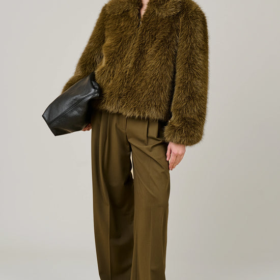 A model wearing a brown shaggy faux fur cropped jacket and matching wide-leg trousers, holding a black leather bag against a neutral gray background.