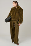 A model wearing a brown shaggy faux fur cropped jacket and matching wide-leg trousers, holding a black leather bag against a neutral gray background.