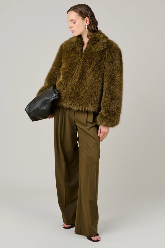 A model wearing a brown shaggy faux fur cropped jacket and matching wide-leg trousers, holding a black leather bag against a neutral gray background.