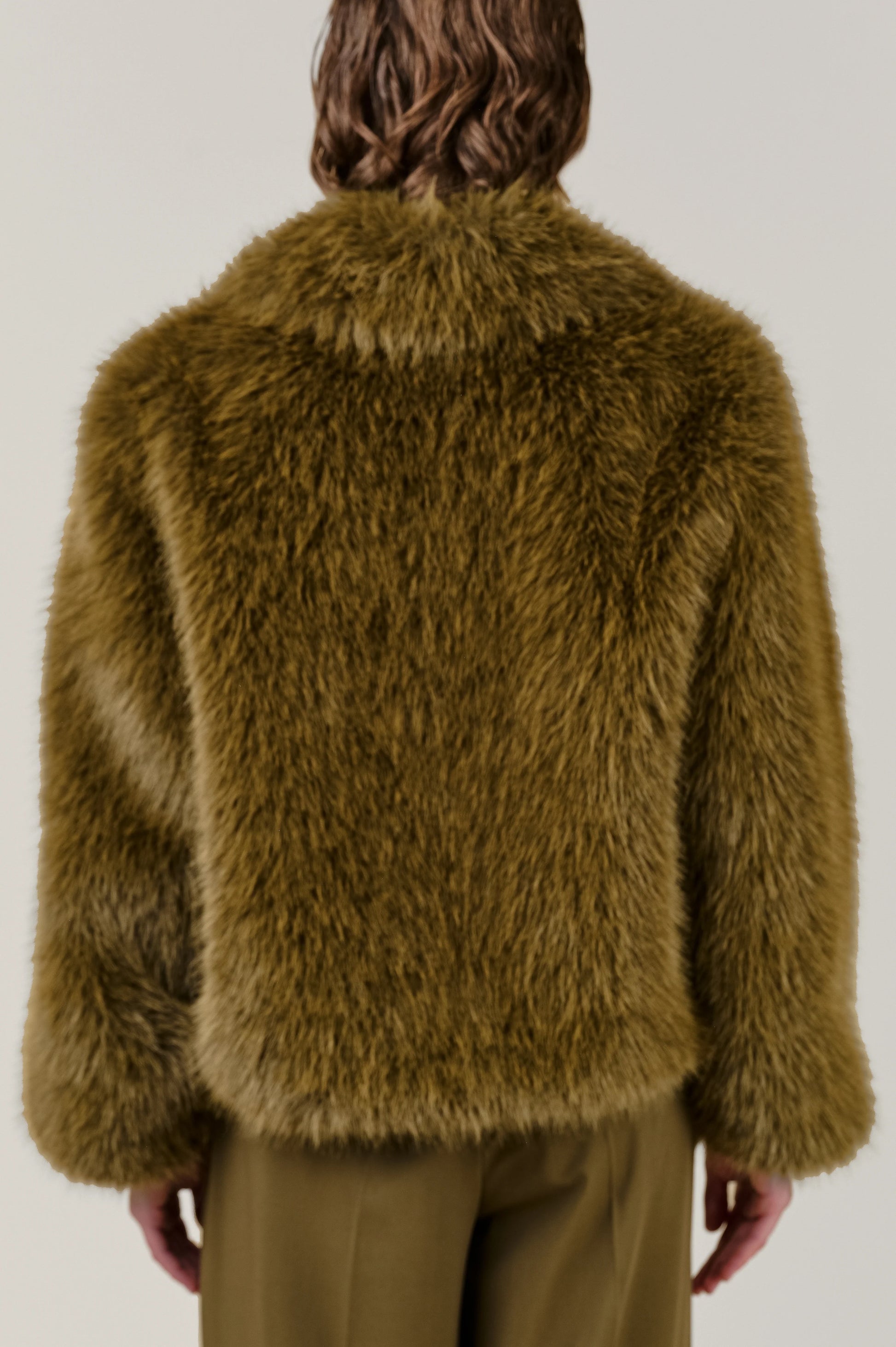 A person viewed from the back wearing a shaggy, textured olive-brown faux fur jacket and matching olive-brown trousers against a plain light gray studio background.