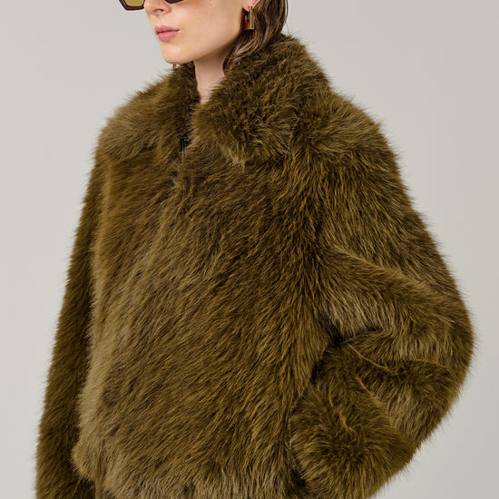 A model wearing a voluminous olive-brown faux fur jacket and matching olive-brown trousers, styled with oversized dark sunglasses, against a solid light gray studio background.