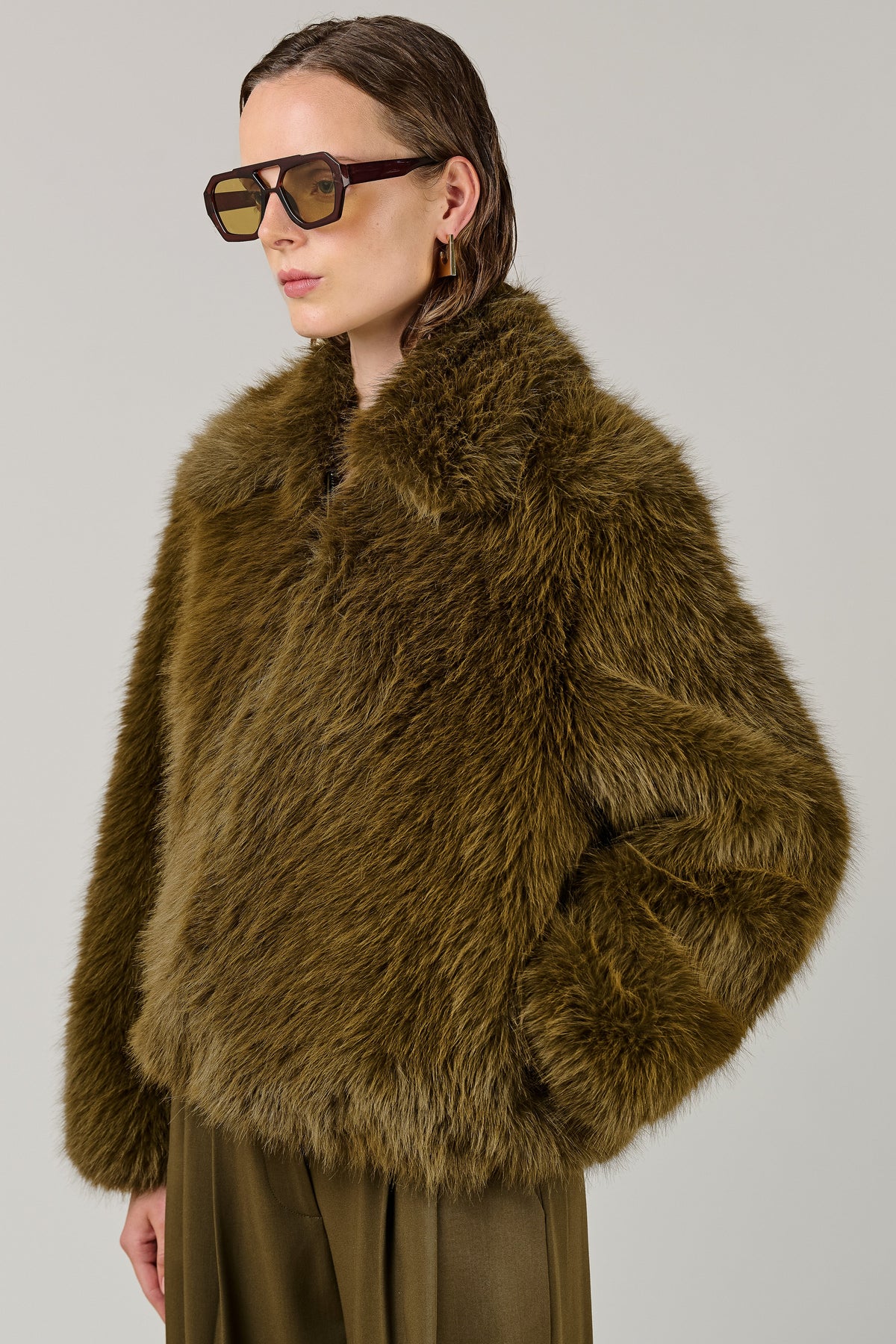 A model wearing a voluminous olive-brown faux fur jacket and matching olive-brown trousers, styled with oversized dark sunglasses, against a solid light gray studio background.