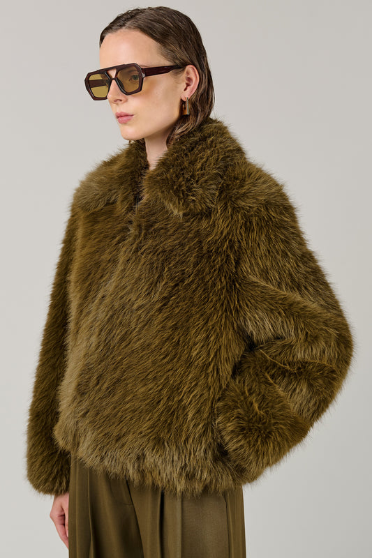 A model wearing a voluminous olive-brown faux fur jacket and matching olive-brown trousers, styled with oversized dark sunglasses, against a solid light gray studio background.