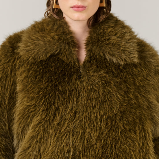 A close-up of a model wearing a voluminous olive-brown faux fur coat with a wide collar, styled with oversized dark sunglasses and gold hoop earrings against a plain light gray background.
