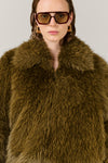 A close-up of a model wearing a voluminous olive-brown faux fur coat with a wide collar, styled with oversized dark sunglasses and gold hoop earrings against a plain light gray background.