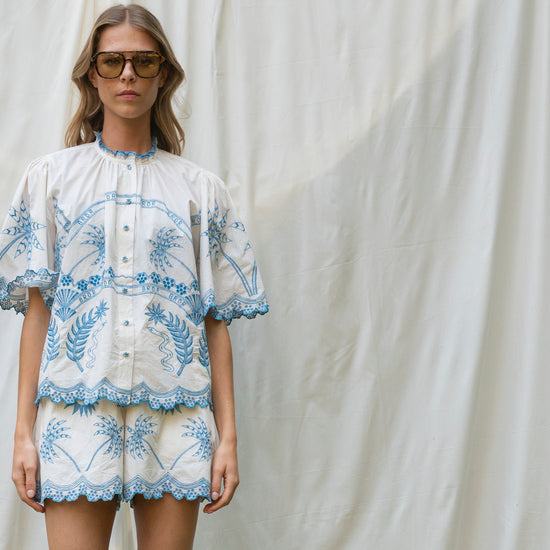 Woman wearing a white blouse with blue floral embroidery against a white curtain.