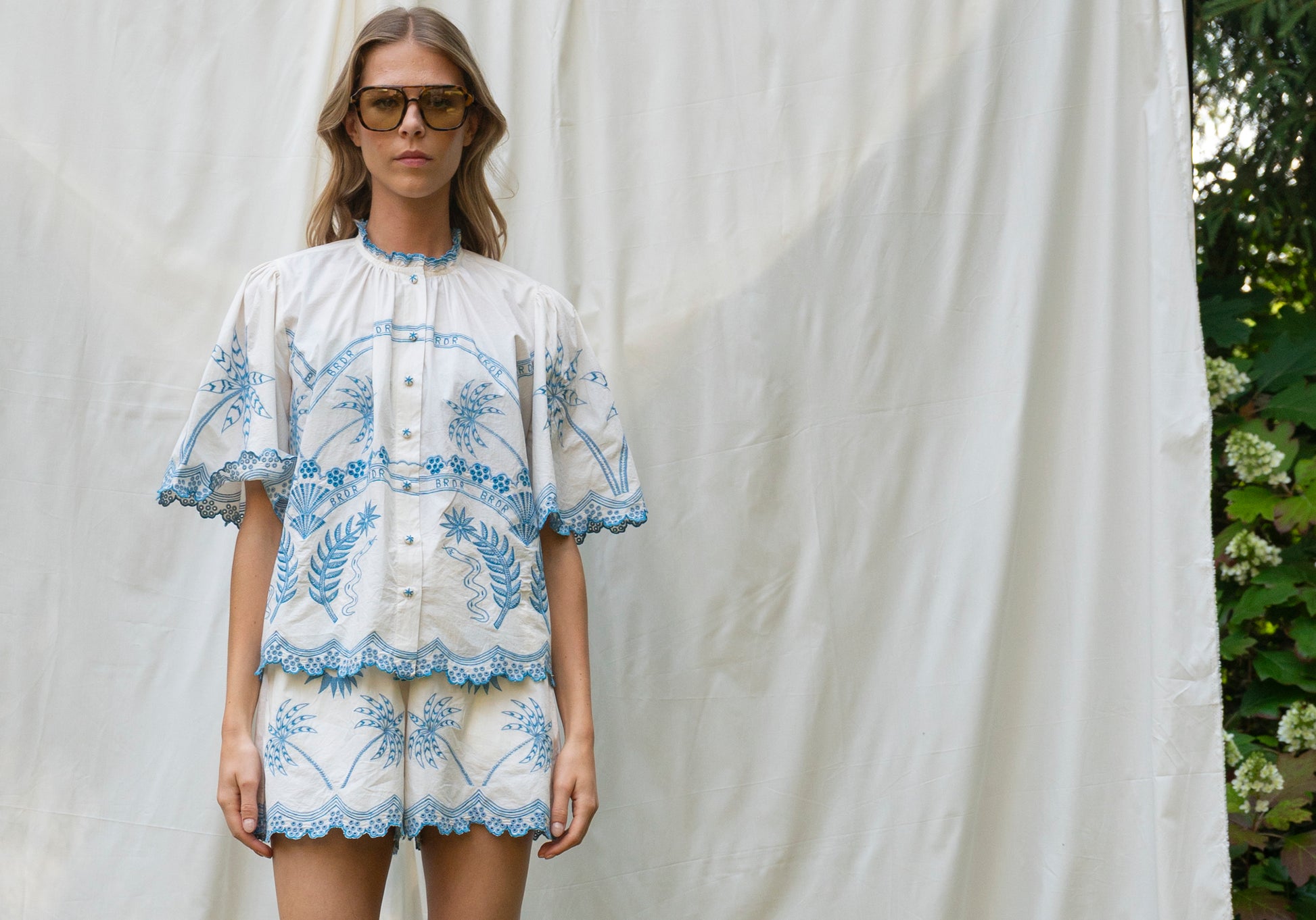Woman wearing a white blouse with blue floral embroidery against a white curtain.