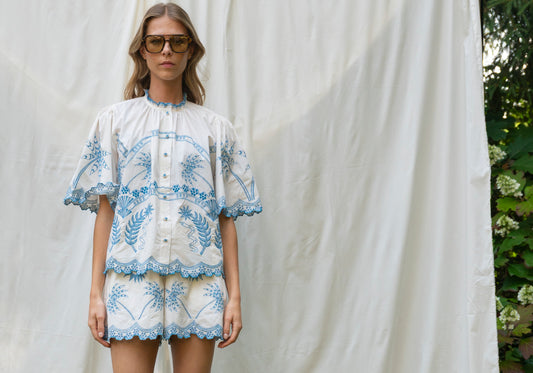 Woman wearing a white blouse with blue floral embroidery against a white curtain.