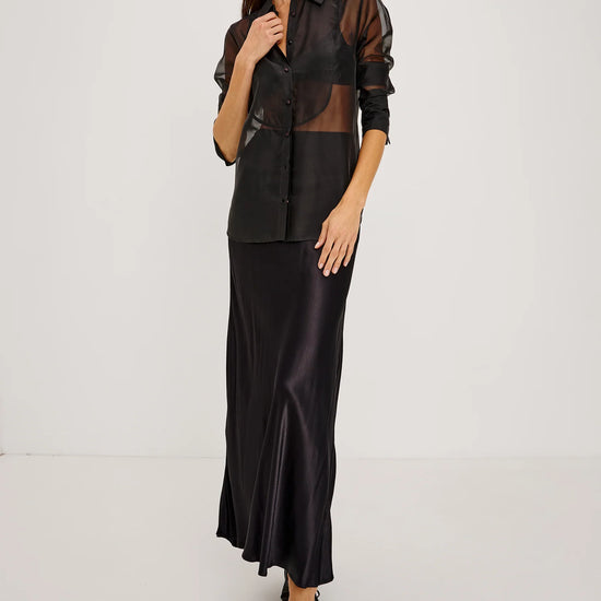 full body shot of a Model wearing a sheer black button up shirt with a black slip skirt and black pointed toe shoes 
