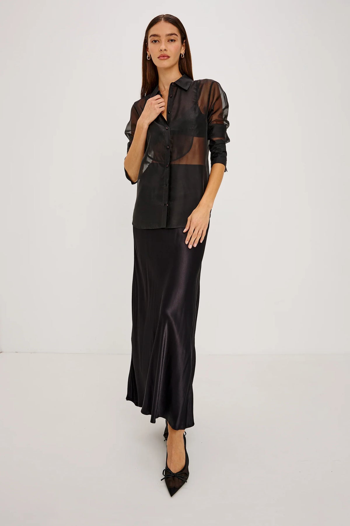 full body shot of a Model wearing a sheer black button up shirt with a black slip skirt and black pointed toe shoes 