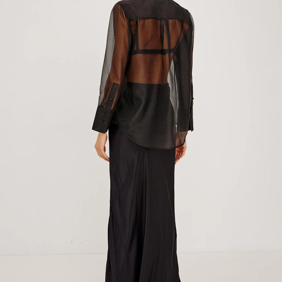 rear shot of a Model wearing a sheer black button up shirt with a black slip skirt  and black heels 