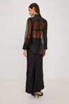 rear shot of a Model wearing a sheer black button up shirt with a black slip skirt  and black heels 