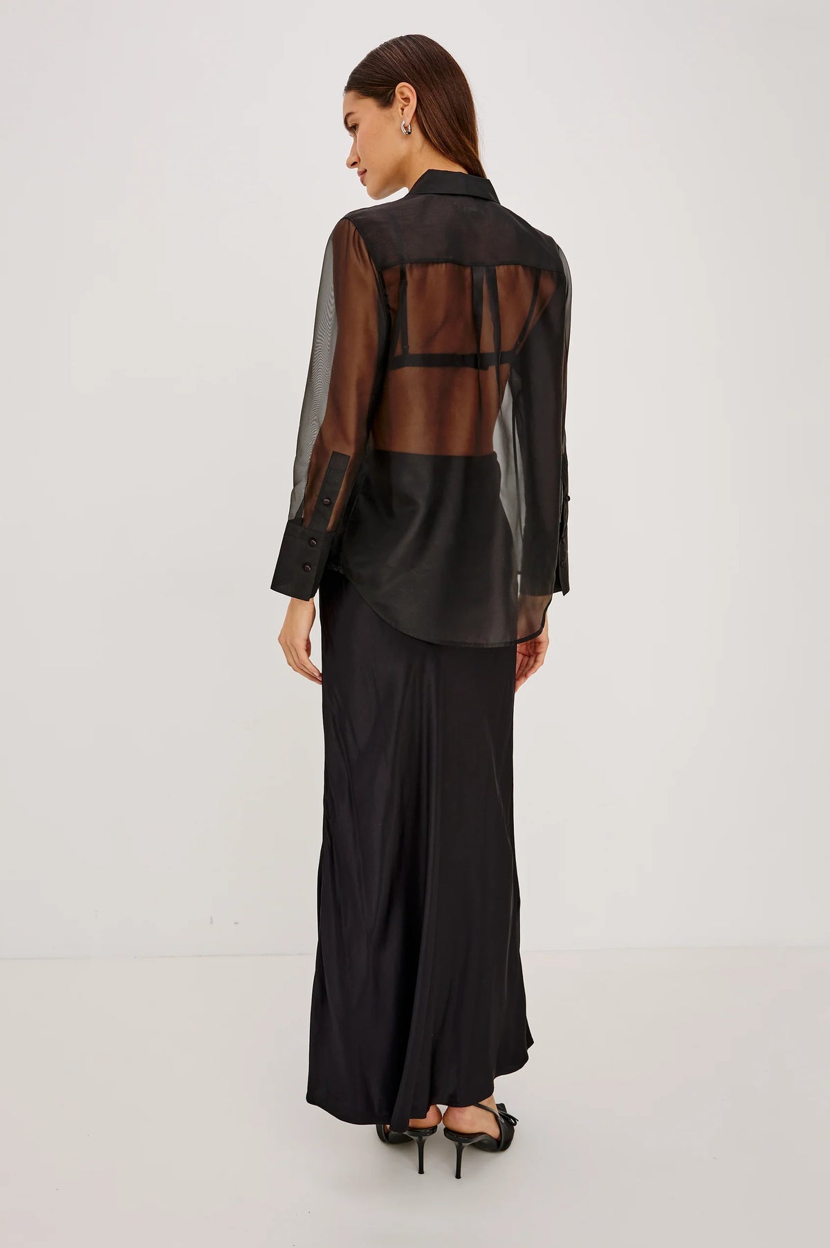 rear shot of a Model wearing a sheer black button up shirt with a black slip skirt  and black heels 