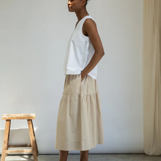 Woman wearing a white top and beige skirt standing in a minimalistic room with a wooden stool and curtain.