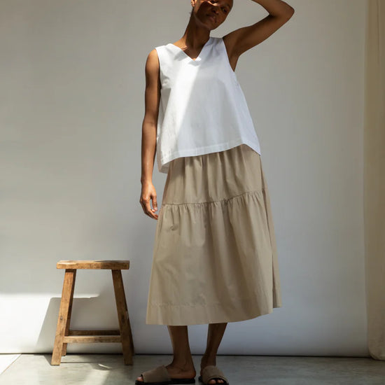 Woman wearing a white sleeveless top and beige skirt standing against a plain wall with a wooden stool.