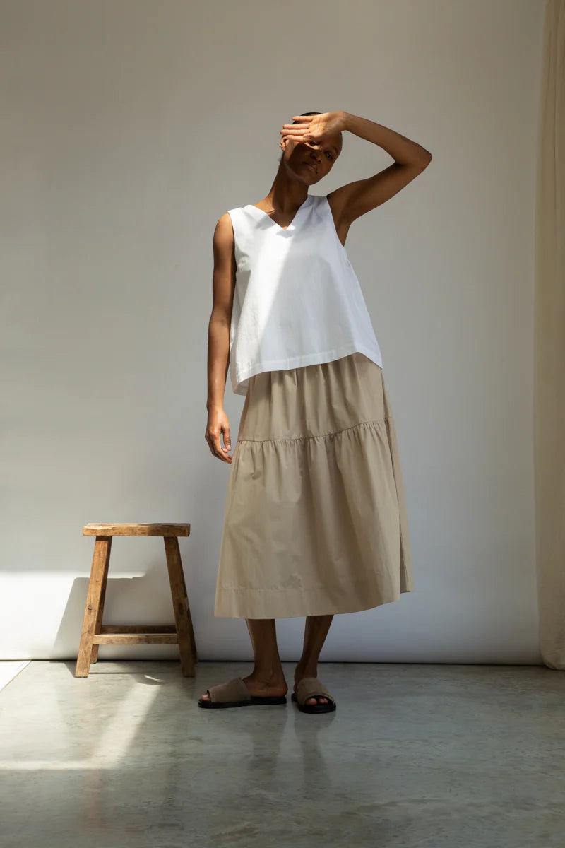 Woman wearing a white sleeveless top and beige skirt standing against a plain wall with a wooden stool.