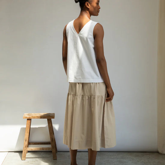 Woman wearing a white sleeveless top and beige skirt standing against a plain wall.